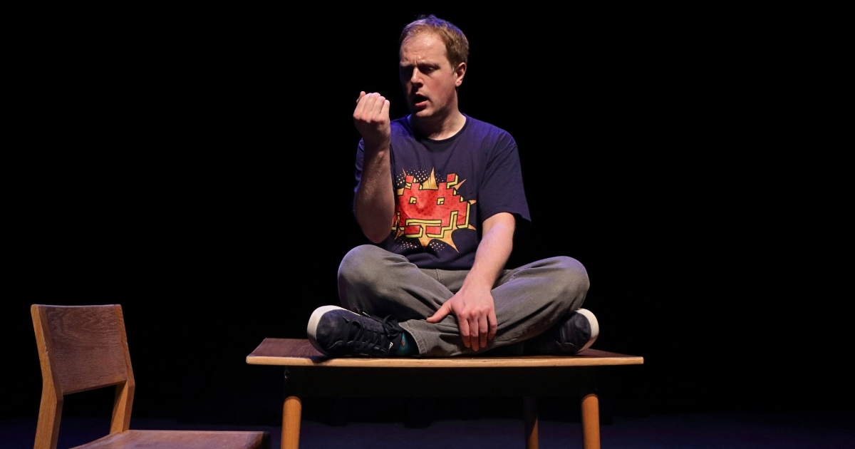 A person sits cross-legged on top of a wooden table and looks at something in the palm of their hand.