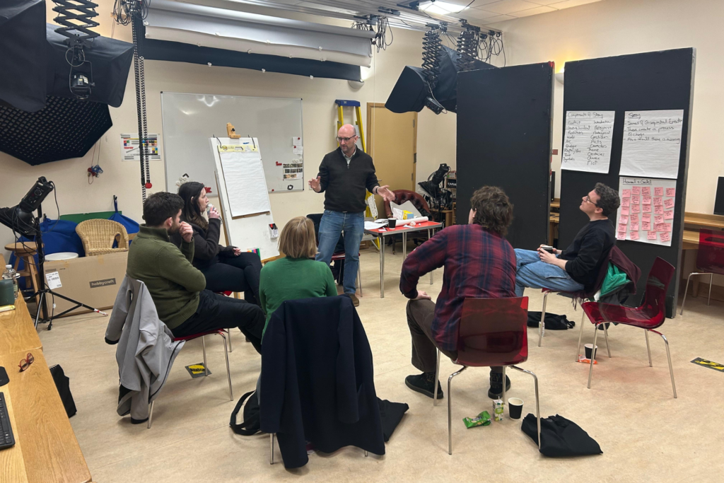 A group of people sit on chairs, facing a person in front of them. Around them hang studio lights.